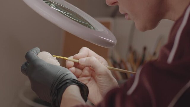 Focused person painting silicone doll head under magnifying lamp using fine brush and gloves, showing precision in detailed artistic creation process