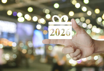 Gift box happy new year 2026 flat icon on finger over blur light and shadow of shopping mall,...