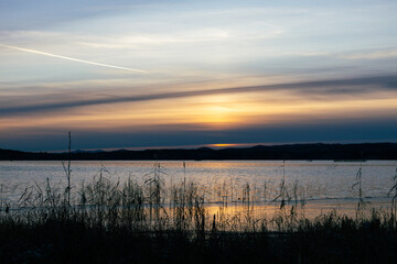 Serene sunset over tranquil lake, with gentle ripples reflecting warm hues of orange and purple, surrounded by distant hills and soft clouds, creating a peaceful natural landscape