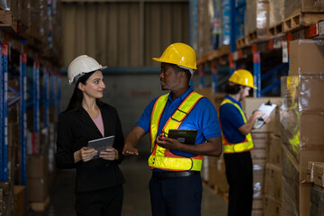 Warehouse worker and female supervisor discussing logistics tasks with digital tablets while another staff inspects inventory. Concept of teamwork, digital operations and smart supply chain