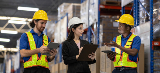 Warehouse workers and female supervisor using digital tablets and clipboard to discuss inventory management. Team collaboration concept in modern logistics and supply chain operations.