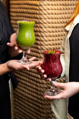 Two friends hold colorful tropical drinks, one green and one red, in a cozy bar. The atmosphere is filled with laughter and the promise of good times, enhancing their joyful moment