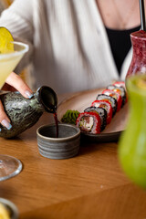 Hand pours rich soy sauce into a small bowl, enhancing colorful sushi rolls on the plate. A refreshing cocktail rests nearby as friends gather for a joyful meal