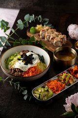 A vibrant bowl of bibimbap garnished with vegetables and a fried egg sits beside sushi rolls and colorful side dishes. The cozy atmosphere is accentuated by green leaves and a touch of elegance