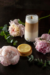 A creamy latte stands tall amidst soft pink peonies and a delicate pastry, inviting moments of calm and enjoyment in a tranquil café atmosphere filled with beauty
