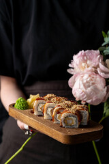 A person holds a wooden platter adorned with beautifully arranged sushi rolls, topped with sesame seeds. Delicate peonies add an elegant touch to the scene, enhancing the culinary experience