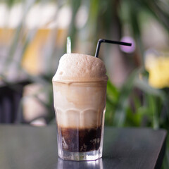 Creamy iced coffee float with thick foam served in a glass on a café table. Soft natural light, blurred green background, and clean composition perfect for beverage marketing, menus, and lifestyle vis