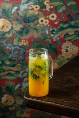 A refreshing cocktail made with vibrant fruits and herbs rests on a rustic wooden table. The background displays a bold floral design that adds character to the setting