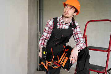 Pensive male laborer, builder or repairman ready for repairing or building, wears overalls, tool belt and protective helmet, works at construction site