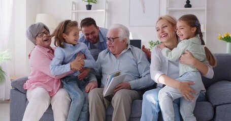 Happy united big multi generation family gathering on sofa together, looking through photo album. Cheerful grandfather showing photographs, sharing joyful memories with children and grandchildren. - Powered by Adobe