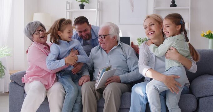 Happy united big multi generation family gathering on sofa together, looking through photo album. Cheerful grandfather showing photographs, sharing joyful memories with children and grandchildren.