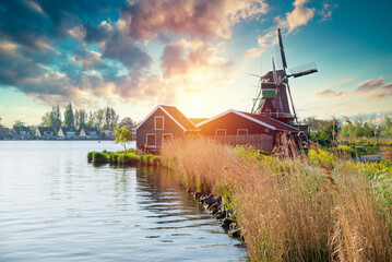 cottage lined waterfront near windmill at dusk with warm rooflines and tall reeds, shimmering water and soft pink sky evoking