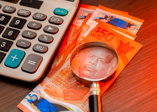An overhead shot of Barbadian 50 dollar banknotes featuring Errol Barrow, scrutinized under a magnifying glass next to a silver calculator on wooden surface highlighting financial auditing and budget.