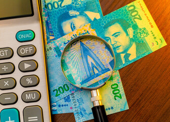 The image features Moroccan 200 Dirham banknotes on a wooden surface, partially viewed through a magnifying glass. A calculator sits to the left, suggesting a theme of finance or auditing.