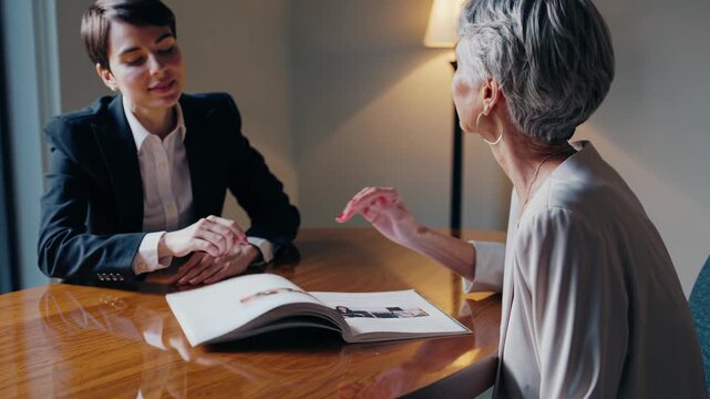 Youthful financial consultant advising an older woman on retirement investments while two businesswomen review a pamphlet and discuss a project at a corporate workstation