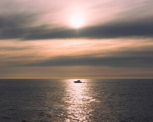 Boat Crossing the Sea at Sunset. A solitary boat sails across the shimmering ocean during a soft, pastel sunset, with warm light reflected on the water under a dramatic sky. 

