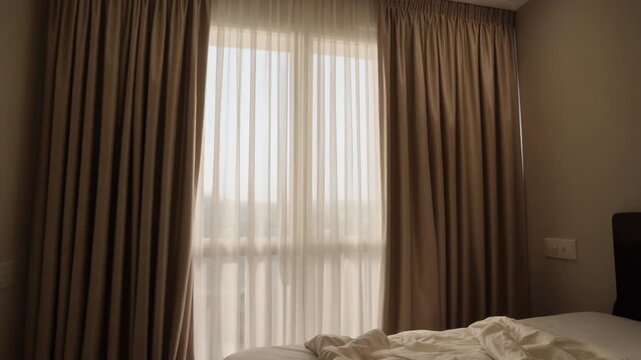A serene bedroom scene viewed from the corner, capturing soft morning light filtering through sheer curtains and brown drapes, with a bed and rumpled bedding.