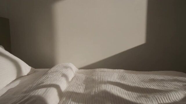 Gentle motion through a minimalist bedroom, fabric textures catching passing light while matte surfaces subtly shift in tone.