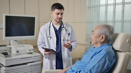 Young doctor in white coat smiling, gesturing with hands while holding tablet, standing next to elderly patient sitting in chair. Medical professional tells aged man about results of tests - Powered by Adobe