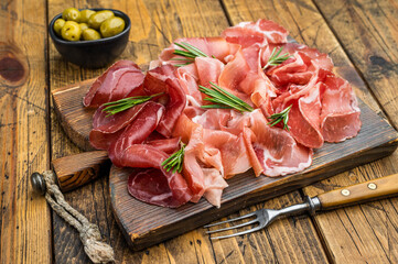Premium dry cured meats on rustic wood: prosciutto, coppa and bresaola garnished with rosemary and green olives, overhead shot.