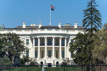 Obraz premium Whitehouse stands in Washington DC under a clear sky with the flag flying and a view of trees nearby during the day