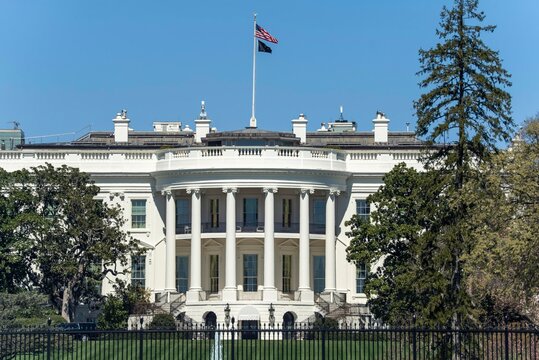 Whitehouse stands in Washington DC under a clear sky with the flag flying and a view of trees nearby during the day