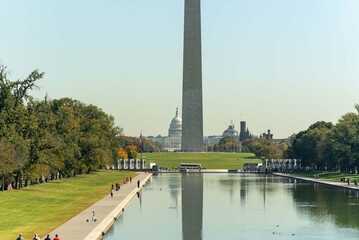 People walk alongside the reflecting pool with the US Capitol and Washington Monument visible in the background on a clear day