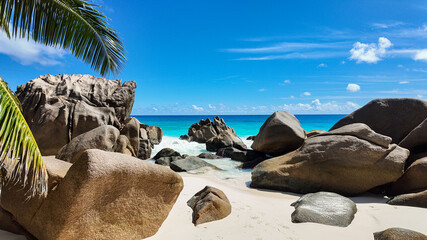 Granite boulders on white sandy beach with turquoise ocean under a blue sky. Palm leaves frame the view. Seychelles, La Digue. Anse Patates. © Alex Traveler