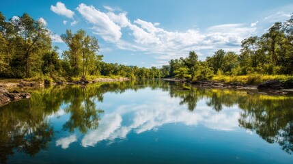 Obraz premium Serene river flowing through a lush green forest under a blue sky with fluffy clouds. Reflection of trees and sky on water.