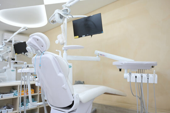 A bright dental operatory with a white treatment chair, multiple instrument trays, overhead lighting, and a wall-mounted monitor