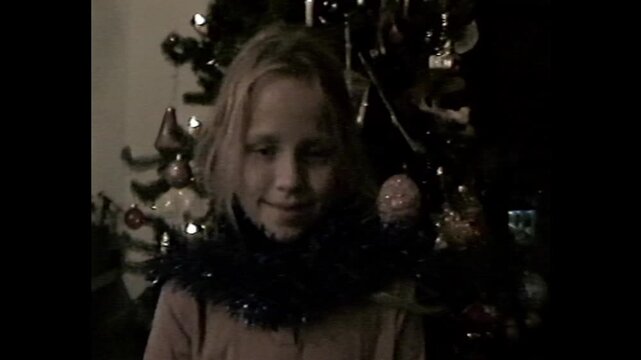 Girl with tinsel wrapped around neck posing near a Christmas tree in retro VHS family footage