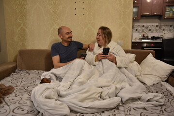 Man and woman sitting together on a bed and holding their smartphones while talking and smiling