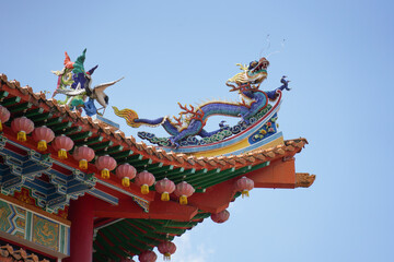 an ornate, traditional Chinese temple with red, gold, and green colors, adorned with pink lanterns, intricate carvings, and a white balustrade under a clear blue sky.