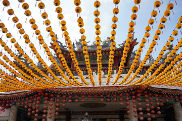 an ornate, traditional Chinese temple with red, gold, and green colors, adorned with pink lanterns, intricate carvings, and a white balustrade under a clear blue sky.