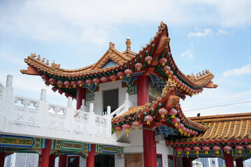 an ornate, traditional Chinese temple with red, gold, and green colors, adorned with pink lanterns, intricate carvings, and a white balustrade under a clear blue sky.