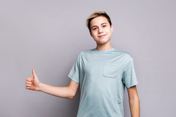 Smiling teenage boy showing thumbs up gesture and looking at camera. Confident preteen male with short blond hair in blue t-shirt posing against gray studio background