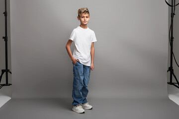Young boy model with stylish haircut standing in studio. Kid modeling new clothing or showcasing child fashion with copy space on gray background