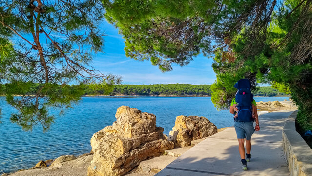 A father hiking with his baby in a carrier walks along a paved coastal path framed by lush pine trees, enjoying scenic blue sea view and active family holiday on a sunny day on Rab Island, Croatia. - Powered by Adobe