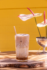 Iced latte served in a textured glass on a wooden table with a vibrant yellow background, capturing modern caf&eacute; aesthetics and trendy summer drink presentation for menus and hospitality marketing.
