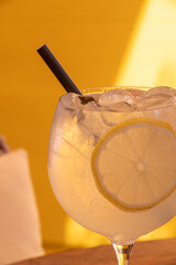 Close-up of a refreshing lemon cocktail with ice in a tall glass against a vibrant yellow background, capturing a bright summer mood ideal for drink menus, bar promotions and hospitality branding.