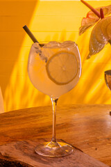 Refreshing lemon cocktail with ice served in a tall glass on a wooden table, lit by warm sunlight with a vibrant yellow background, perfect for summer menus, bar promotions and drink advertising.