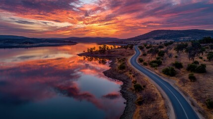 Fototapeta premium A beautiful sunset over a lake with a road in the distance. The sky is filled with orange and pink hues, creating a serene and peaceful atmosphere. The water reflects the colors of the sky
