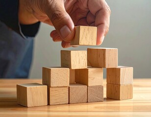 A hand places a wooden block atop a pyramid of similar blocks, on a light-wood surface