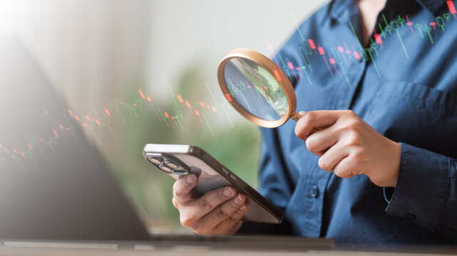 A person analyzing financial charts using a smartphone and magnifying glass at a workspace. Finance concept. Stock market trader chart. Finance lifestyle concept. - Powered by Adobe