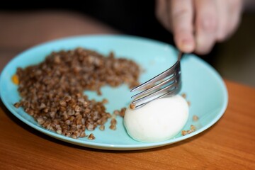 Metal fork cutting boiled egg on plate with buckwheat porridge during breakfast