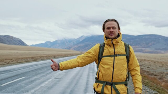 Smiling young man in a yellow jacket with a backpack trying to stop a car with a thumb-up gesture while hitchhiking on an empty mountain road during his adventure travel