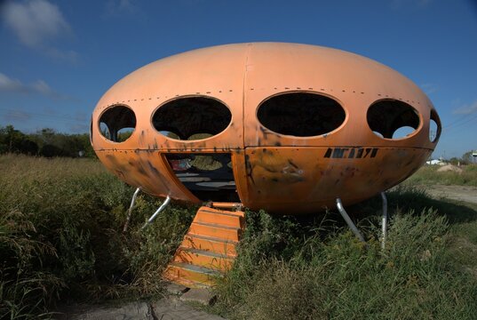 An abandoned ufo shaped house sits in an overgrown field. 