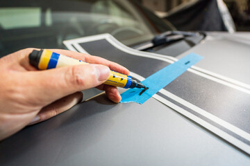 Worker installing stickers on a car in a garage, close-up on a hand that holds a marker and makes marks for sticking. Car decal application on the hood.
