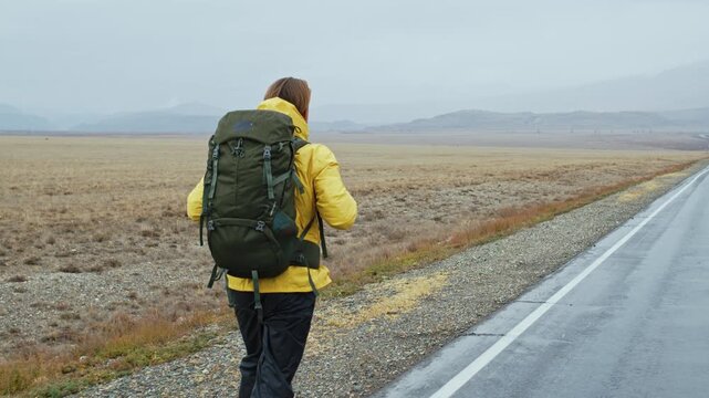 Man wearing a yellow jacket and carrying a large backpack hiking alone on the side of a desolate road. Rear view of a male tourist trekking through a vast, empty landscape on a foggy day