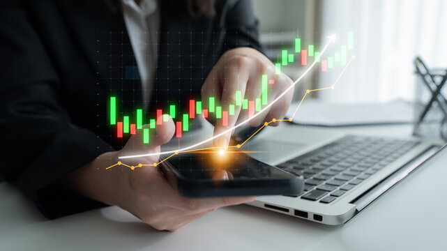 A business professional analyzes stock market data using a smartphone, displaying a graphical candlestick chart alongside a laptop in a modern office setting. Impute
